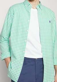 Zeleno-bílá kostkovaná košile na knofl íky z bavlny, s ležérním límcem a malým fialovým logem na hrudi. Nenošena přes bílou košili.