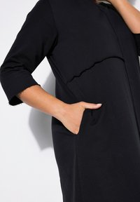 Vestido negro con mangas tres cuartos, que presenta un bolsillo frontal, bordes deshilachados y un corte relajado. Fabricado con una tela suave y elástica.