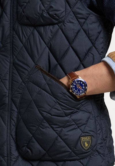 Marineblå quilted vest med diamantmønster, prydet med en brun kordfløjpocket trim og et gult broderet logo. Et rundt blåt ur med brun læderrem bæres på håndleddet.