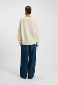 Kremowy sweter dzianinowy o oversize'owym kroju, z prążkowanymi mankietami i hemem, w zestawieniu z szerokimi, ciemnoniebieskimi dżinsami. Miękka tekstura, prosty design.