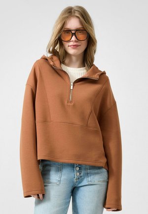Jonge vrouw draagt een bruine zip-up sweatshirt, witte trui eronder, lichtblauwe spijkerbroek en grote amberkleurige bril, staand tegen een grijze achtergrond.