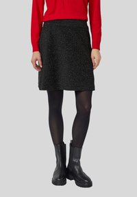 Eine Frau trägt einen glitzernden schwarzen Rock, schwarze Strumpfhosen, schwarze Stiefeletten und ein rotes Langarmoberteil, während sie vor einem grauen Hintergrund steht.