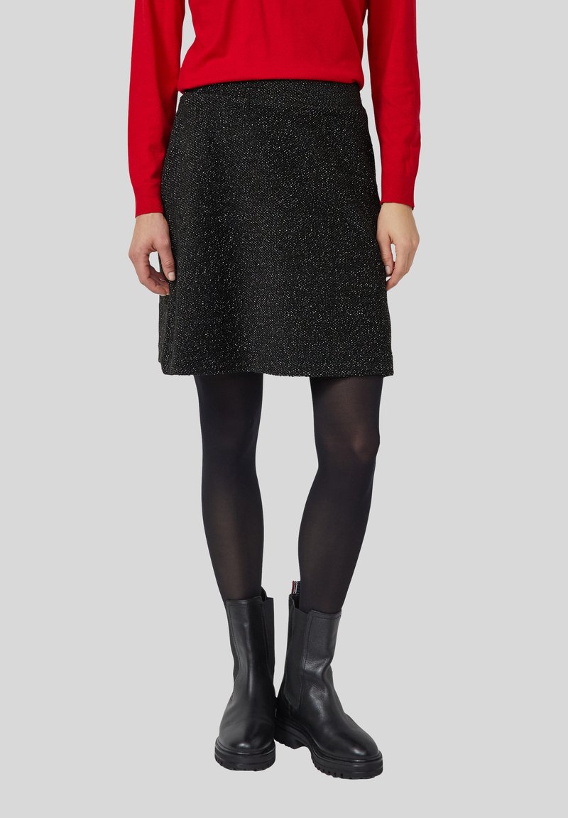 Eine Frau trägt einen glitzernden schwarzen Rock, schwarze Strumpfhosen, schwarze Stiefeletten und ein rotes Langarmoberteil, während sie vor einem grauen Hintergrund steht.