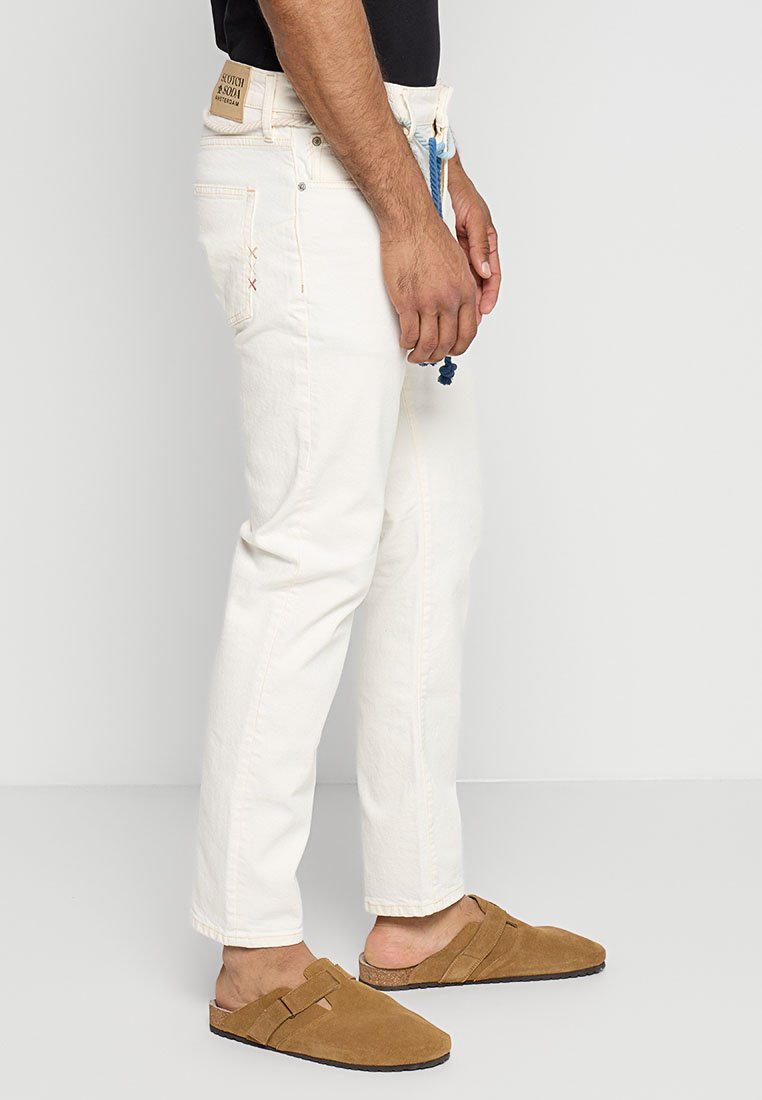 Jeans blancos de corte recto, con costuras contrastantes, un cordón azul y una etiqueta en la parte trasera de la cinturilla. Combinados con zapatos marrones de tipo slip-on.