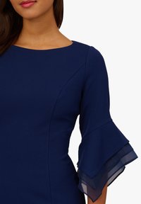 Navyblaues Kleid mit Bateau-Ausschnitt und weit ausgestellten Glockenärmeln mit透明em Lagenakzent. Glatter Stoff mit strukturierten Nähten.