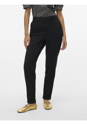Frau trägt schwarze, maßgeschneiderte Hosen mit aufgekrempelten Säumen, goldene Ballerinas und ein schwarz-weiß gestreiftes Kurzarmshirt vor einem schlichten Hintergrund.