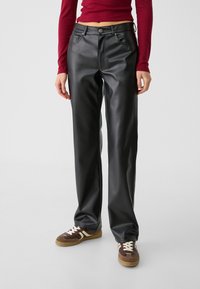 Černé kalhoty z faux kůže se střihovým střihem rovného nohavic, s předními kapsami a zapínáním na knoflík, doplněné hnědými teniskami.