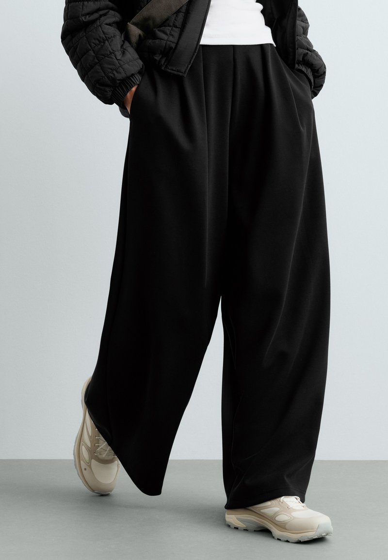 Personne portant un pantalon large noir ample, des baskets blanches, une veste matelassée noire, les mains dans les poches, marchant sur un sol gris.