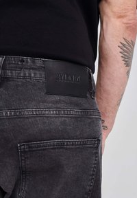 Juodos džinsinės kelnės su odiniu užrašu "2YDNA" ant juosmens. Yra užpakaliniai kišeniai ir tekstūruotas audinys. Matomo nusidėvėjimo nėra.