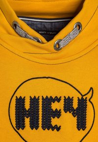 Felpa gialla con la scritta "HEY" ricamata in blu navy all'interno di un fumetto. Corda grigia con occhielli metallici attorno al cappuccio.