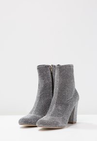 Botas de tobillo grises con un acabado texturizado y brillante. Cuenta con un cierre de cremallera y un tacón alto grueso. Diseño de punta redondeada.