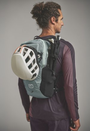 Man with curly hair in a ponytail wearing a purple long-sleeve shirt and carrying a light blue backpack with a white helmet and trekking poles attached.