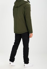 Vihreä oliivinvärinen toppatakki hupulla, jossa on vetoketjukiinnitys ja sivutaskut, yhdistettynä mustiin slim fit -housuihin ja valkoisiin nilkkureihin.