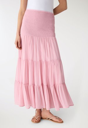 Donna che indossa una gonna lunga rosa a vita alta con balze e cinturino arricciato in vita e sandali beige, in piedi davanti a uno sfondo semplice.