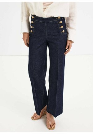 Dunkelblaue Jeans mit weitem Bein und hoher Taille, vorne an den Taschen mit zwei Reihen goldener Knöpfe, kombiniert mit einem weißen Hemd und beigen Sandalen.