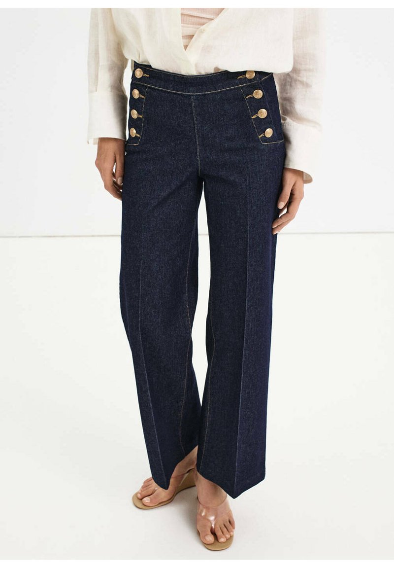 Jean large bleu foncé taille haute avec deux rangées de boutons dorés sur les poches avant, porté avec une chemise blanche et des sandales beiges.