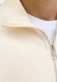 Beige zip-jacka med en texturerad krage och silverdetaljer på dragkedjan, lagd över en vit skjorta. Blankt tyg med modern design.
