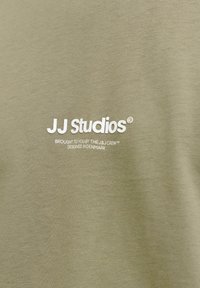 Olivengrønn bomulls-T-skjorte med hvit tekst: "JJ Studios" og "Brought to you by The J&J Crew™ Designed in Denmark." Myk tekstur.
