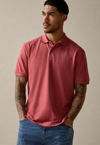 Polo de manga corta en rosa apagado, con cuello clásico y logo bordado. Combinado con jeans azules, mostrando un ajuste relajado y material de algodón.