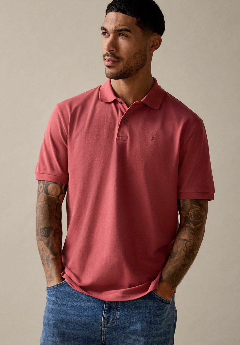 Polo de manga corta en rosa apagado, con cuello clásico y logo bordado. Combinado con jeans azules, mostrando un ajuste relajado y material de algodón.