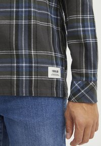 Koszula flanelowa w kratę w kolorze czarnym, zielonym i niebieskim, wykonana z miękkiego materiału, z metką z nazwą marki umieszczoną na dolnym lewym szwie.
