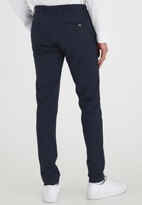 Pantaloni slim-fit blu navy con una texture liscia, caratterizzati da due tasche posteriori con bottoni e dettagli sottili, abbinati a sneakers bianche.