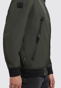 Maslinasto zeleni bomber jakna s glatkom površinom, rebrastim manžetama i džepom na patent zatvaraču na rukavu. Ima logo oznaku na ramenu.