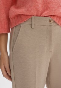 Khaki-Hosen aus strukturiertem Stoff, mit flachem Frontdesign und Seitentaschen. Kombiniert mit einem weichen, korallfarbenen Strickpullover, der gerippte Akzente zeigt.