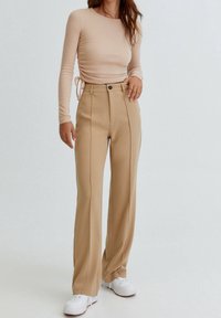 Beżowa bluzka z długim rękawem i marszczeniem po bokach. Beżowe spodnie o wysokim stanie, proste nogawki, z przeszyciami i zapięciem na guziki z przodu. Białe sneakersy.