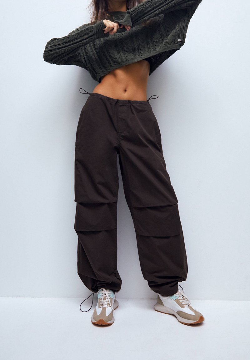 Bruine, baggy broeken met plooien en een taille met trekkoord, gecombineerd met een cropped, structuurdonkergroen sweater en lichte sneakers met accenten.