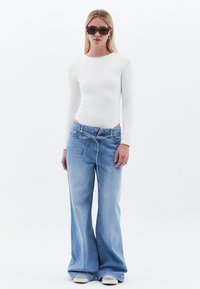 Weißer Langarm-Body, kombiniert mit hellblauen weit geschnittenen Jeans. Die Jeans haben einen Kordelzugbund und eine lockere Passform. Weiße Sneaker vervollständigen den Look.