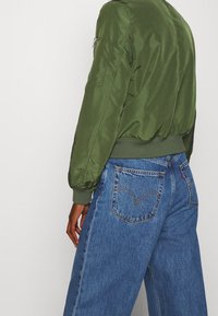 Zielona satynowa kurtka bomberka z prążkowanymi mankietami i krawędzią, noszona na niebieskich jeansach Levi's z wysokim stanem, posiadających klasyczny tylny wzór kieszeni.