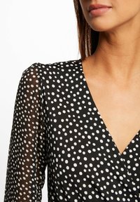 Blusa negra con lunares blancos, que presenta un escote en V y mangas largas transparentes. La tela parece suave y ligera, adecuada para usar en capas.