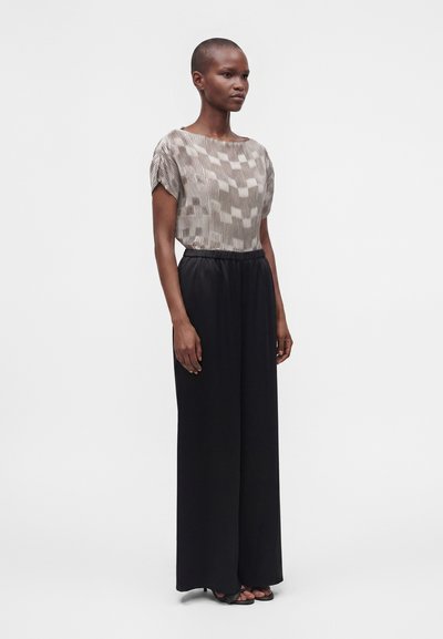 Femme debout portant un haut à manches courtes à motifs rentré dans un pantalon noir taille haute à jambes larges et des sandales noires sur un fond uni.
