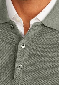 Graues Strick-Poloshirt mit strukturiertem Muster, ausgestattet mit einer drei-Knopf-Leiste und einem weißen Hemd mit Kragen darunter.