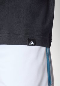 T-shirt en coton noir avec une étiquette du logo Adidas au bas, associé à un short blanc avec des rayures latérales bleues et une texture de tissu lisse.