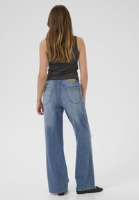 Donna con lunghi capelli castani, indossa un top senza maniche nero, jeans blu larghi e scarpe nere basse, in piedi con le spalle rivolte verso l'osservatore su uno sfondo bianco.