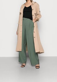 Beige trenchcoat med knappar, svart ribbad linne, skräddarsydda gröna vida byxor och svarta klacksandaler. Slät tyg, elegant passform.