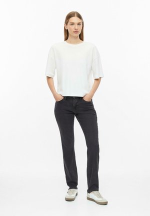 Vrouw staat met handen in de zakken, draagt een wit korte mouwen top, zwarte spijkerbroek en witte sneakers op een witte achtergrond.