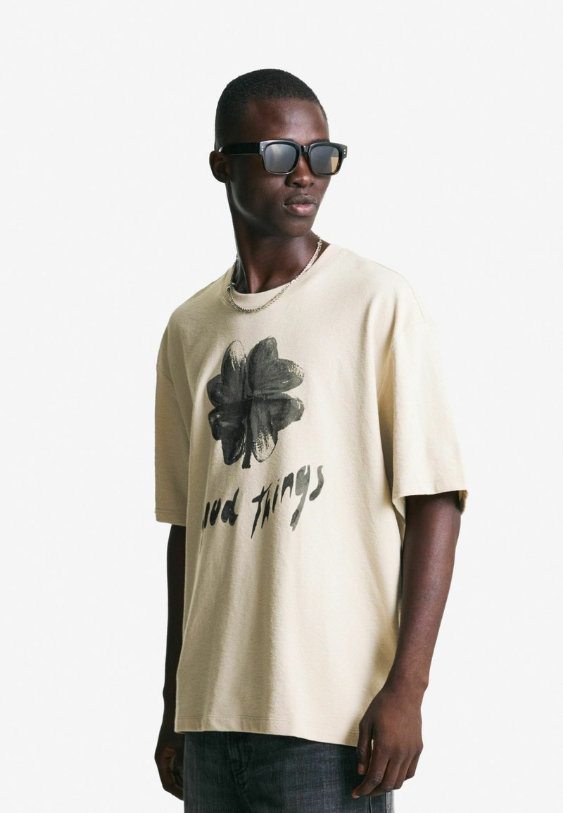 Bež majica oversized s crnom grafiko deteljice in napisom "dobre stvari". V kombinaciji s sivimi kavbojkami in temnimi sončnimi očali, v stoječem položaju.