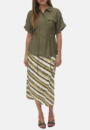 Femme portant une chemise boutonnée à manches courtes de couleur olive avec des liens à la taille, associée à une jupe midi rayée blanche et olive, et des talons noirs.