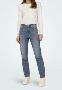 Lehká krémová košile s dlouhým rukávem v kombinaci s modrými džíny s vysokým pasem, rovného střihu a jemným vyblednutím. Nošeno s bílými teniskami.