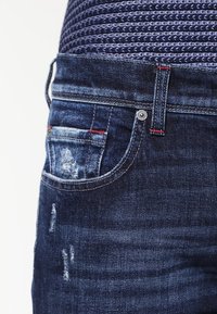 Gros plan sur un jean en denim bleu foncé avec des détails usés et des couture rouges près de la poche avant et de la passant de ceinture.