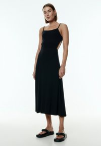 Schwarzes Midi-Kleid mit tailliertem Oberteil, Spaghettiträgern und offenen Seiten. Hergestellt aus einem glatten, dehnbaren Stoff. Getragen mit schwarzen Sandalen.