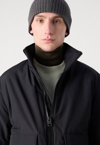 Jachetă neagră cu fermoar, cu guler înalt, asociată cu un pulover verde închis cu guler înalt. Modelul poartă o căciulă gri cu textură în relief. Material fin și design minimalist.