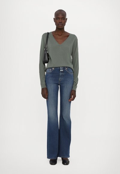 Suéter verde de cuello en V, de mangas largas, combinado con unos vaqueros de campana azules. Textura suave, ajustado en la cintura, mostrando una silueta simple. Bolso negro.