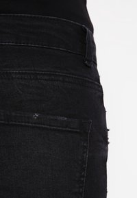 Jeans de denim negro con un diseño ajustado, costuras visibles y un parche desgastado en el bolsillo trasero, con una cintura de altura media.