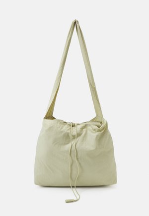 Sac bandoulière en tissu beige clair avec longue bandoulière et fermeture par cordon suspendu sur un fond uni.