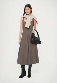 Jauna moteris vilki be rankovių rudu languotu sukniu, juodais batais, ant pečių užsirišusi kreminės spalvos megztinį, laiko juodą odinę rankinę.