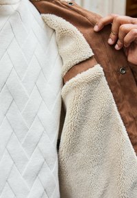 Jachetă din suede maro cu căptușeală din fleece crem, evidențiind un interior texturat și un exterior neted, parțial descheiat pentru a dezvălui straturile.
